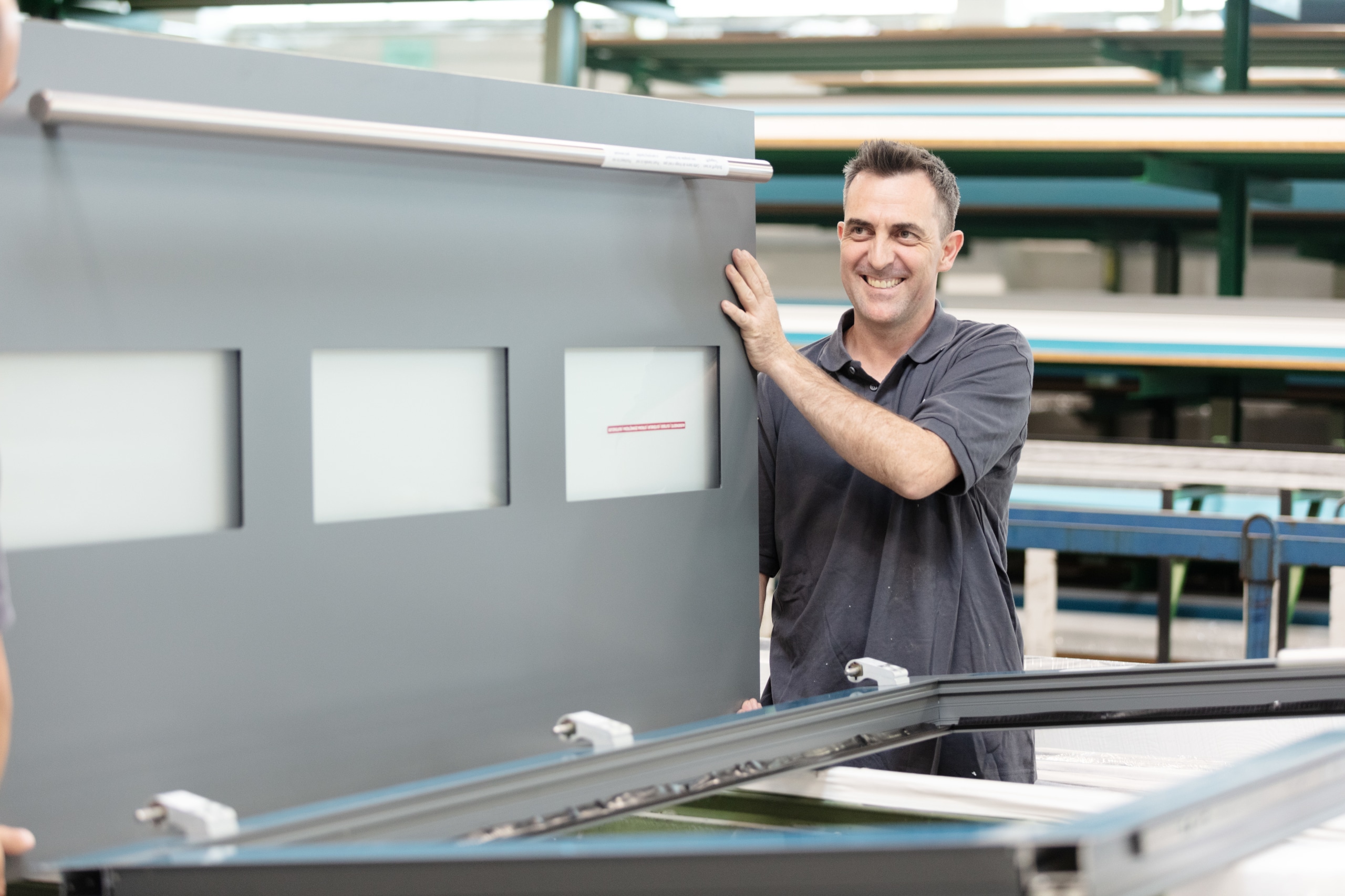 Ein l&auml;chelnder Mann in einer Werkstatt hebt eine gro&szlig;e graue Metallplatte mit drei rechteckigen Fenstern an.