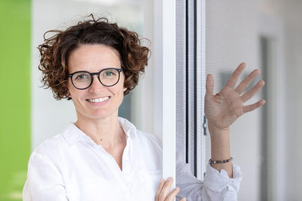 Ein l&auml;chelnder Servicemitarbeiter (m/w/d) im Innendienst mit lockigem Haar und Brille, der ein wei&szlig;es Hemd tr&auml;gt, steht am Fenster und winkt.