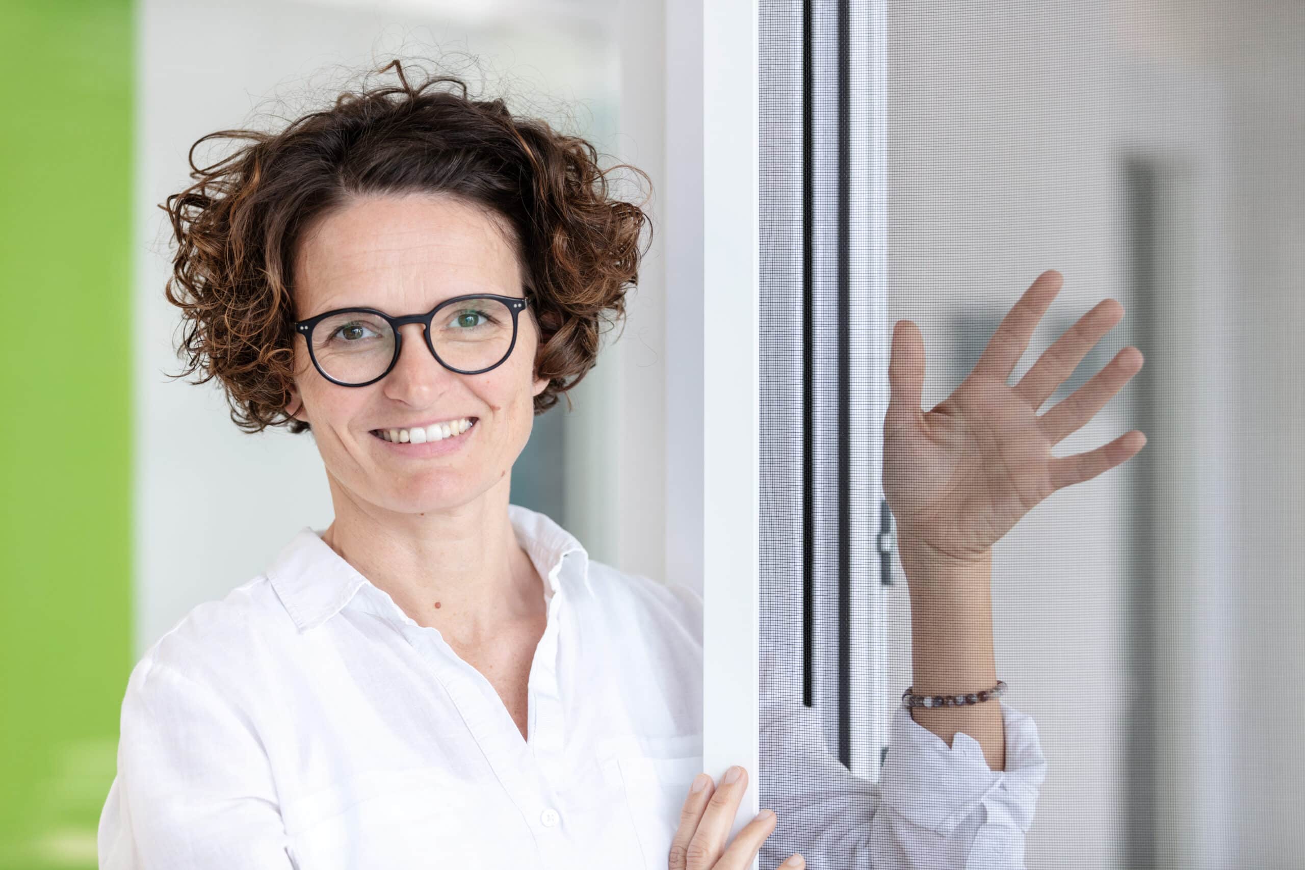 Ein l&auml;chelnder Servicemitarbeiter (m/w/d) im Innendienst mit lockigem Haar und Brille, der ein wei&szlig;es Hemd tr&auml;gt, steht am Fenster und winkt.