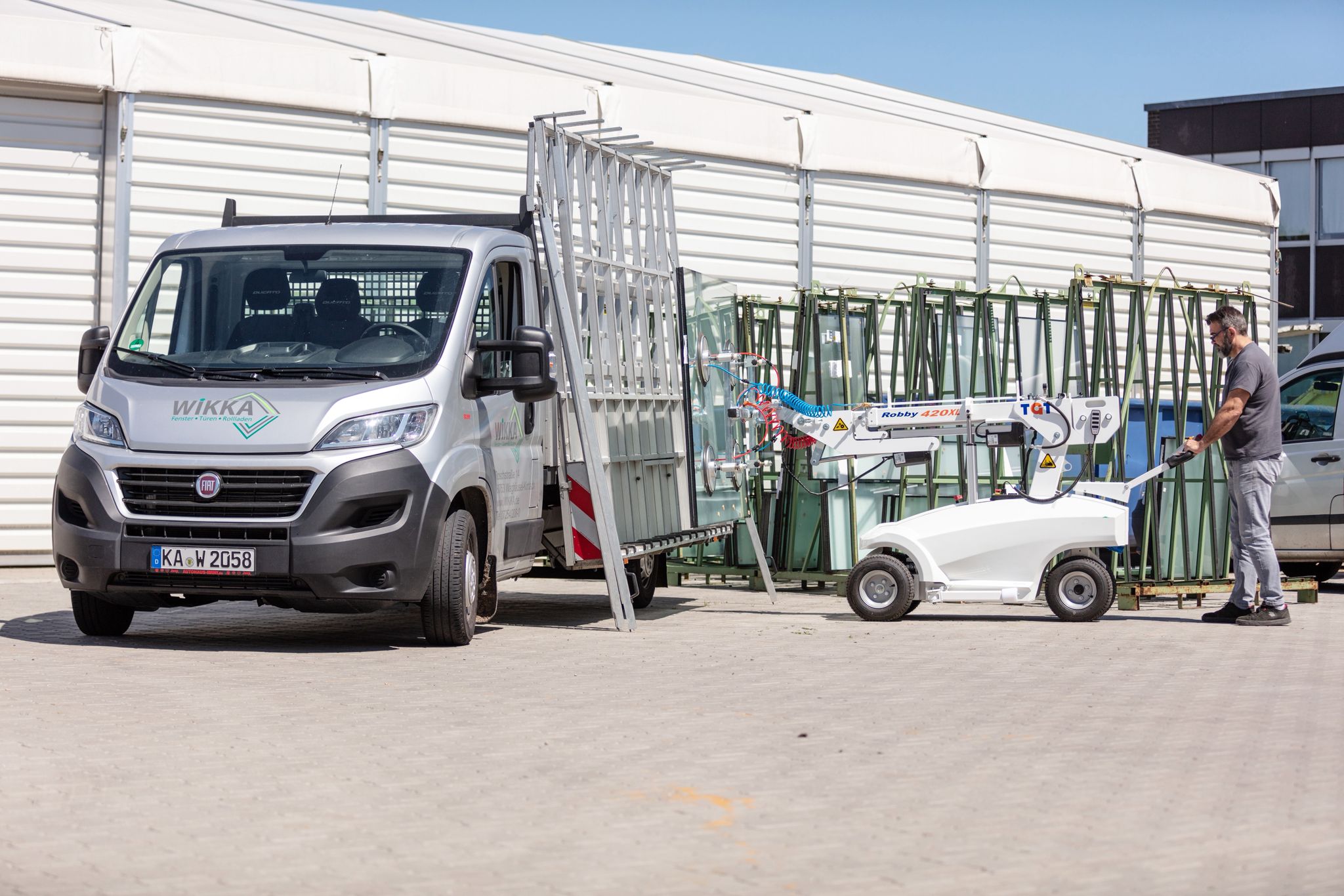 Ein Monteur / Servicetechniker (m/w/d) in Festanstellung nutzt eine kleine Hebeb&uuml;hne, um eine Glasscheibe in die N&auml;he eines Lieferwagens zu transportieren, der neben einem Lagergeb&auml;ude geparkt ist.